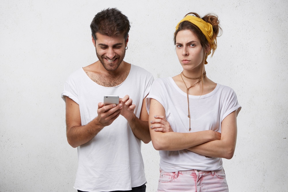 Frau und Mann stehen nebeneinander, er scrollt am Smartphone un sie blickt skeptisch in die Kamera - Symbolbild für den Ratgeber: Wann ist Eifersucht berechtigt?