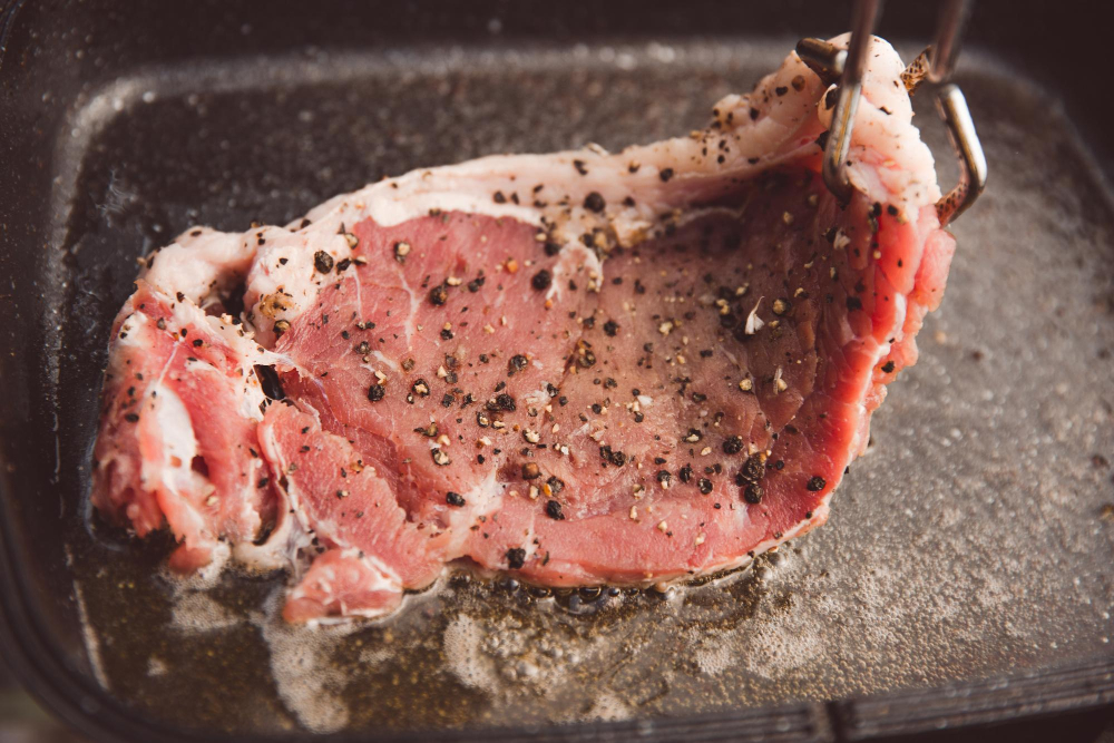 Ein Stück Fleisch, welches mit einer Zange in die Pfanne gelegt wird als Symbolbild für den Ratgeber: Fleisch lässt Wasser beim Anbraten