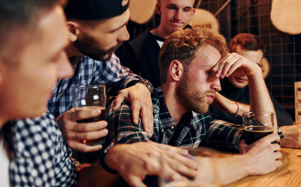 Junge Männer an der Bar versammeln sich um einen Mann, der sich nciht so gut fühlt als Symbolbild für den Ratgeber: Schnell wieder nüchtern werden