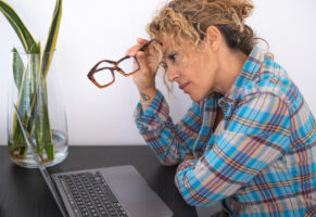 Frau mit lockigem Haar, die ihre Brille am Bügel mit zwei Fingern festhält und genervt auf den Laptop starrt / Symbolbild: Schlechte Verbindung: Was hilft Repeater oder Mesh?