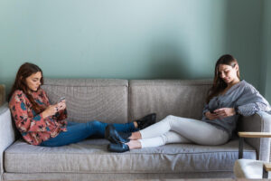 Zwei junge Frauen die beide gegenüber auf dem Sofa liegen und am Smartphone scrollen als Symbolbild für den Ratgeber: 100 Tipps gegen Langeweile