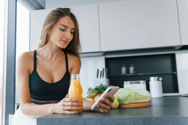 Junge Frau steht an einer Kücheninsel, ist am Smartphone und trinkt Orangensaft, neben hier ist frisches Gemüse als Symbolbild für den Ratgeber: Vor oder nach dem Sport essen