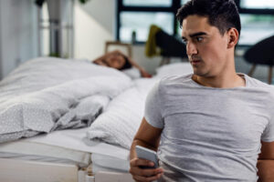 Mann sitzt ruhelos am Ende des Bettes während seine Frau schläft als Symbolbild für den Ratgeber: Typisches Verhalten nach dem Fremdgehen