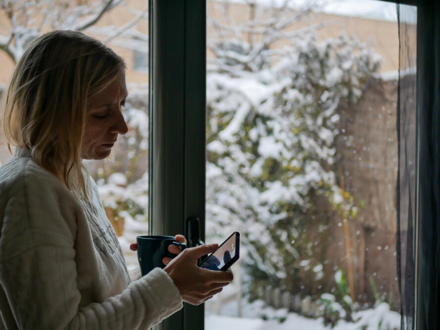 Frau steht am Fenster mit Smartphone und Tee in der Hand, draußen ist es verschneit als Symbolbild für den Ratgeber: Richtig lüften bei Kälte