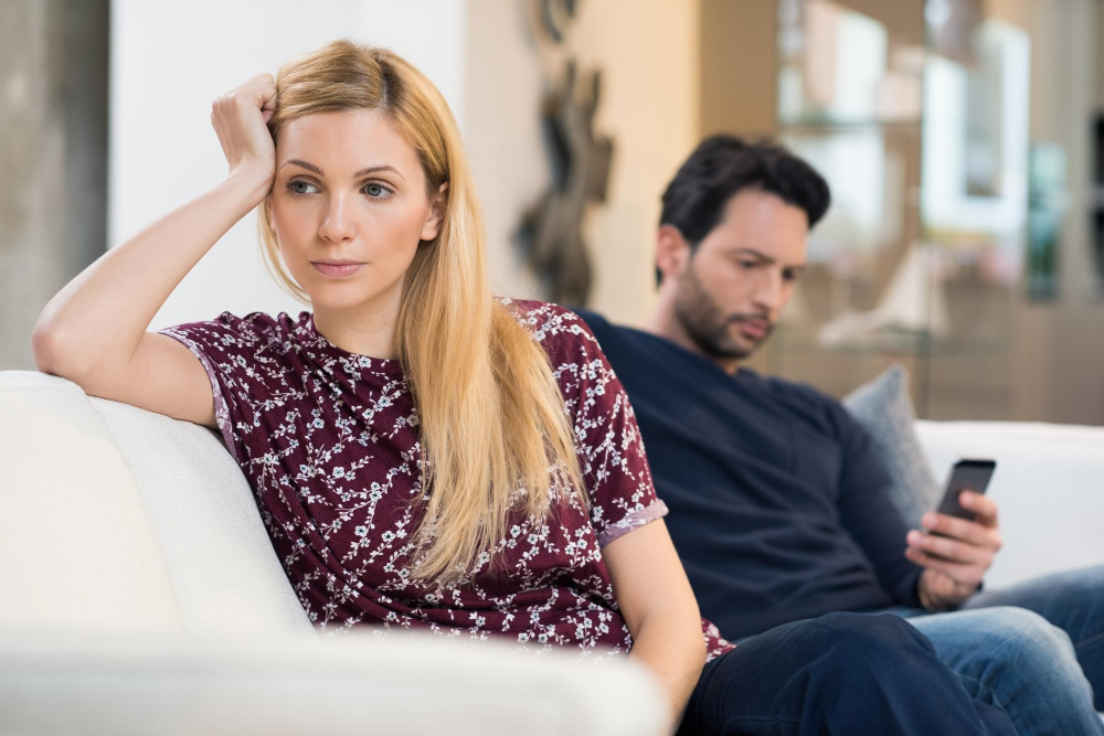 Ein Paar sitzt auf der Couch, der Mann scrollt unaufmerksam durchs Handy während die Frau gedankenversunken in die Luft starrt als Symbolbild für den Ratgeber: Vermisst ein verheirateter Mann seine Affäre?