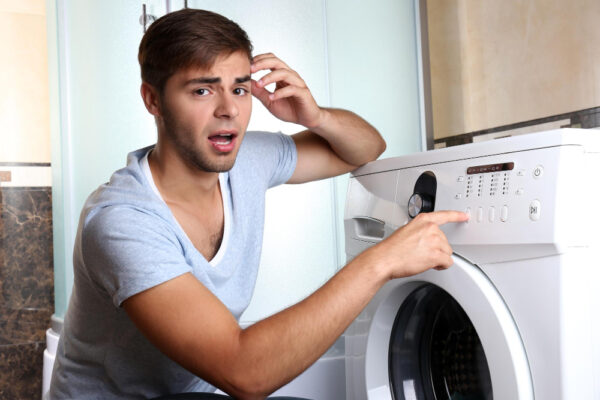 Junger Mann hockt vor einer Waschmaschine und ist fassungslos als Symbolbild für den Ratgeber: Waschmaschine riecht verbrannt