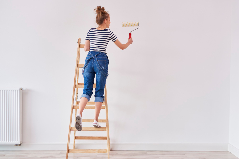 Junge Frau steht auf der Leiter und streicht die Wand als Symbolbild für den Ratgeber: Wandfarbe aus Kleidung entfernen