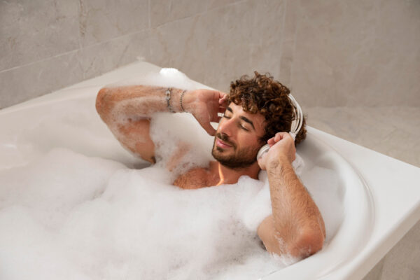 Junger Mann entspannt sich von Schaum bedeckt in einer Badewanne und hört Musik mit Kopfhörern als Symbolbild für den Beitrag: Wie viel Liter passen in eine Badewanne?