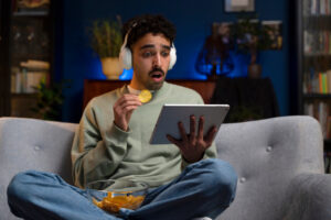 Junger Mann sitzt auf der Couch und sieht auf sein Tablet mit Snacks in der Hand als Symbolbild für den Ratgeber: Streaming-Dienste kostenlos
