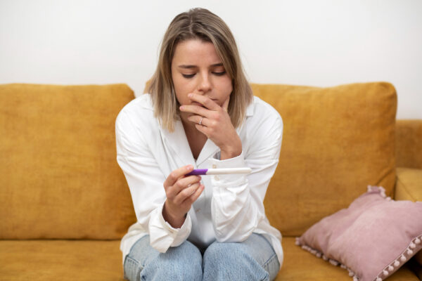 Eine Junge Frau auf einem Sofa die nervös auf ihren Schwangerschaftstest schaut als Symbolbild für Ratgeber: Kann man schwanger werden, wenn man seine Tage hat