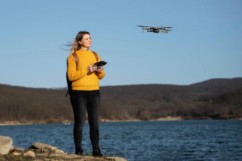 Junge Frau steht am Ufer und steuert eine Drohne als Symbolbild für den Ratgeber: Muss man Drohnen registrieren?