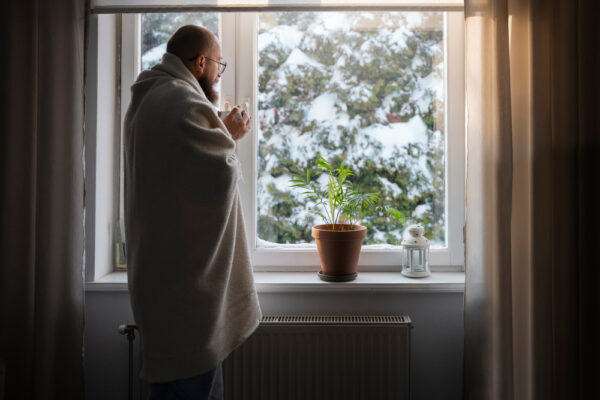 Mann steht in Decke gemummelt vor dem Fenster und blickt hinaus in eine Schneelandschaft als Symbolbild für den Ratgeber: Fenster auf Winter umstellen