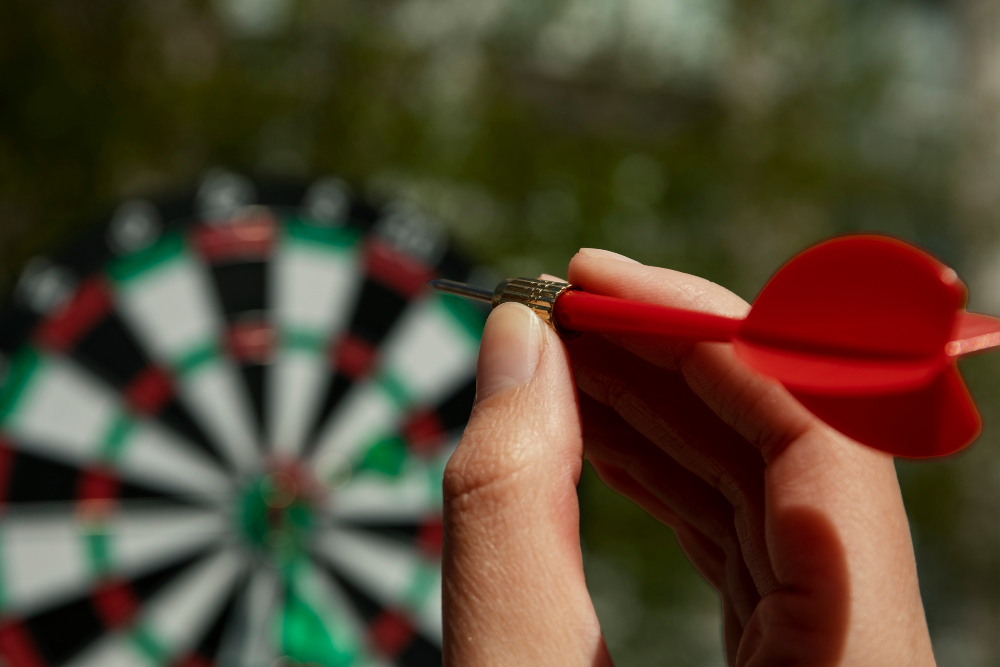 Hand zielt mit rotem Dartpfeil auf eine Dartscheibe als Symbolbild für den Ratgeber: Dart Abstand zur Scheibe