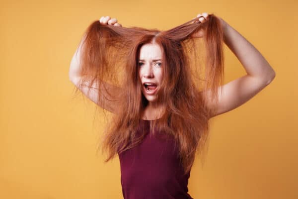 Junge Frau mit roten Haaren hebt ihr Deckhaar und ist entsetzt als Symbolbild für den Ratgeber: Strohige Haare