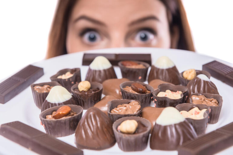 Junge Frau blickt mit weit aufgerissenen Augen hinter einer Platter voller Pralinen und Konfekt hervor als Symbolbild für den Ratgeber: Heißhunger auf Süßes