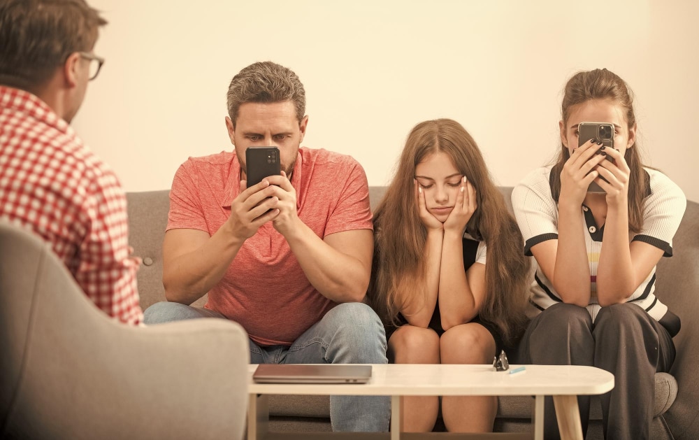 Mutter, Vater, Tochter und Therapeut sitzen an einem Tisch, die Eltern haben aber beide das Smartphone vor der Nase während die Tochter schmollt als Symbolbild für den Ratgeber: Kommunikationsprobleme in der Familie lösen