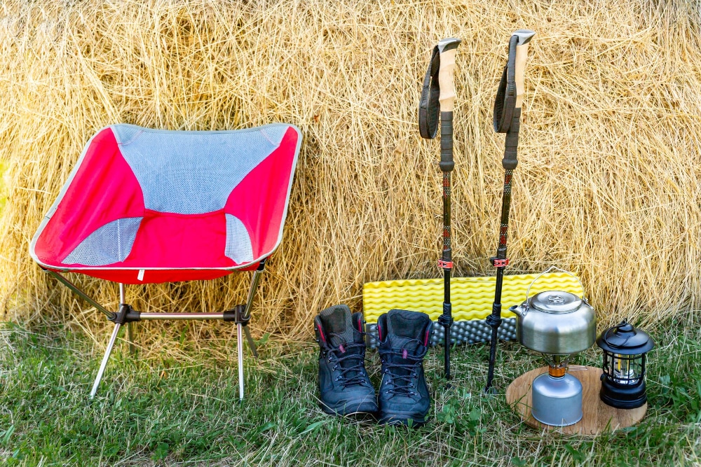 Faltbarer rot-grauer Campingstuhl, Wanderstiefel, Isomatte, Gaskocher, Öllampe und Waderstöcke als Teil der Camping Checkliste