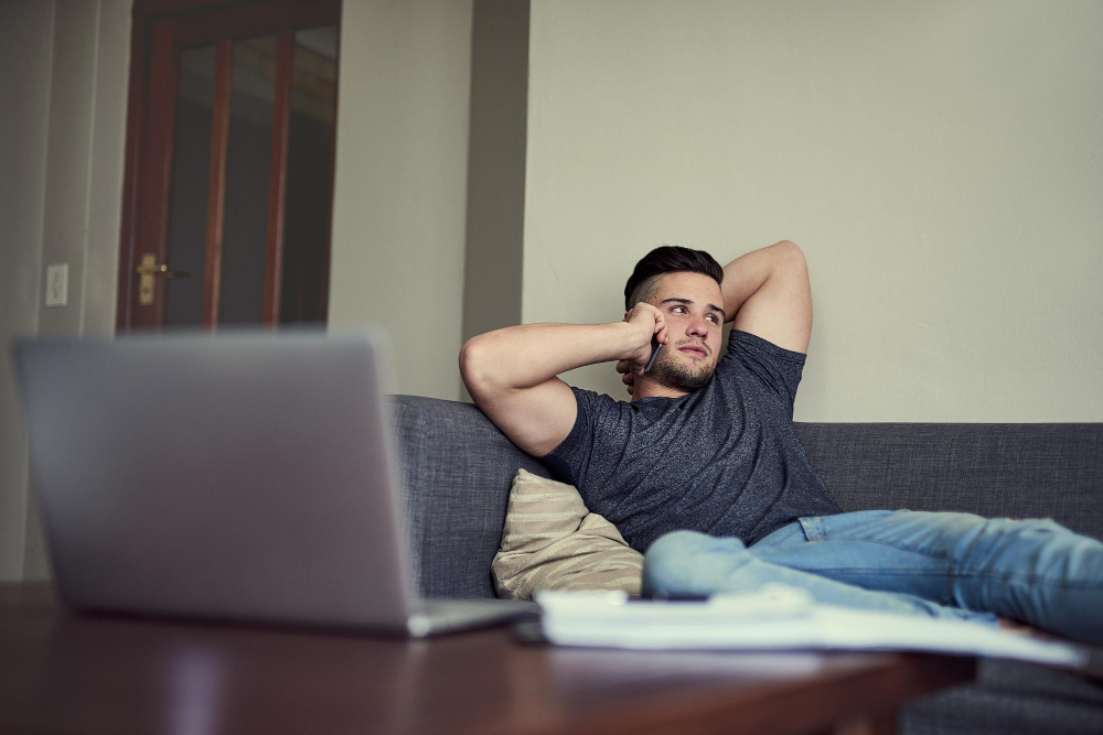 Junger Mann liegt auf der Couch und telefoniert mit dem Smartphone vor einem Laptop als Symbolbild für den Ratgeber: PayPal-Konto im Minus