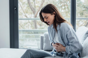 Junge Frau sitzt auf dem Sofa und hält sich den Bauch als Symbolbild für den Ratgeber: Übelkeit nach dem Essen?