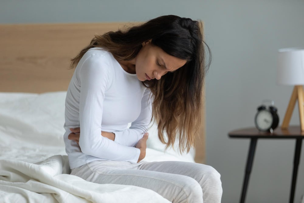 Junge Frau sitzt auf dem Bett und hält sich quälend den Bauch / Sybolbild für Unterleibsschmerzen