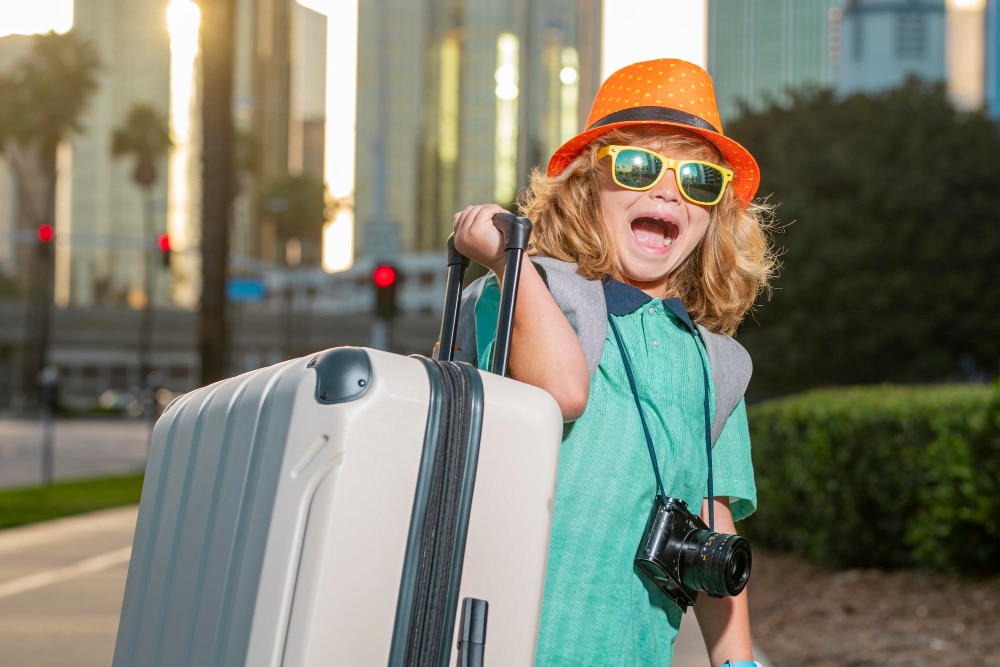 Grinsendes Mädchen mit Sonnenbrille euphorisch mit viel zu großem Koffer als Symbolbild für den Ratgeber: Die besten Reiseziele mit Kindern