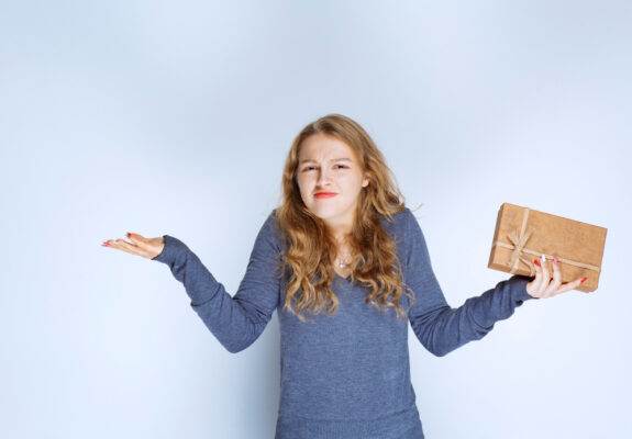 Eine Frau mit einem Geschenk in der Hand hebt fragend beide Arme (symbolisch für den Ratgeber: Geschenke für Leute, die nichts wollen)