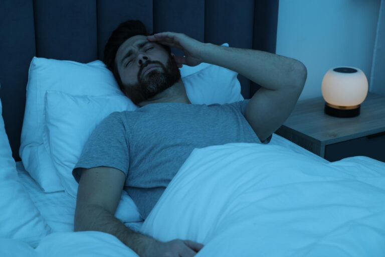 Mann liegt im Bett mit einem besorgten Gesichtsausdruck und fasst sich an die Stirn, symbolisch für das Thema, zu früh aufzuwachen.