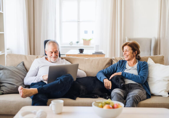 Senioren sitzen ganz bequem auf der Couch wobei der Mann am Laptop Eingaben macht, symbolisch für Onlinebanking