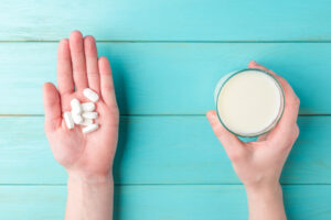 Eine Hand hält mehrere weiße Tabletten, während die andere Hand ein Glas Milch hält, auf einem blauen Holzuntergrund. Das Bild veranschaulicht das Thema 'Antibiotika und Milch' und verdeutlicht die mögliche Wechselwirkung zwischen Medikamenten und Milchprodukten.