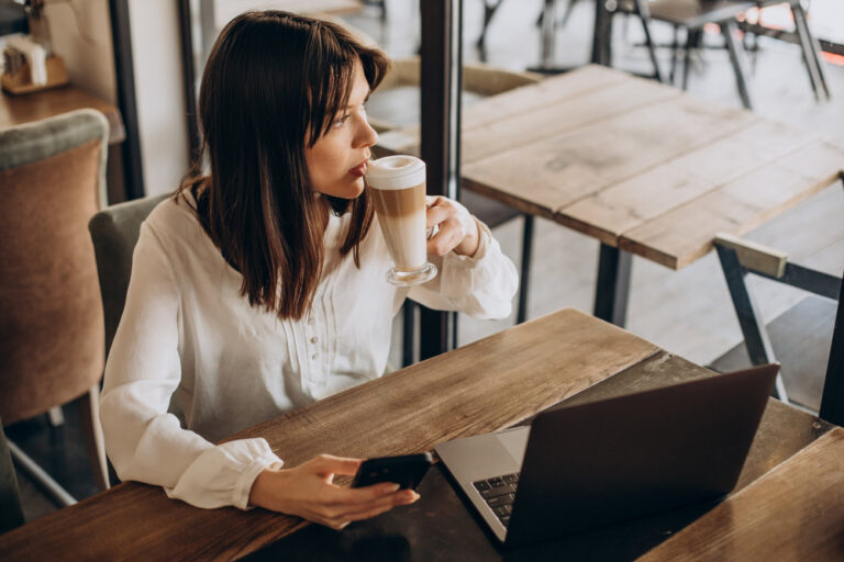 Frau trinkt Latte Macchiato im Café und arbeitet mit Laptop und Smartphone.