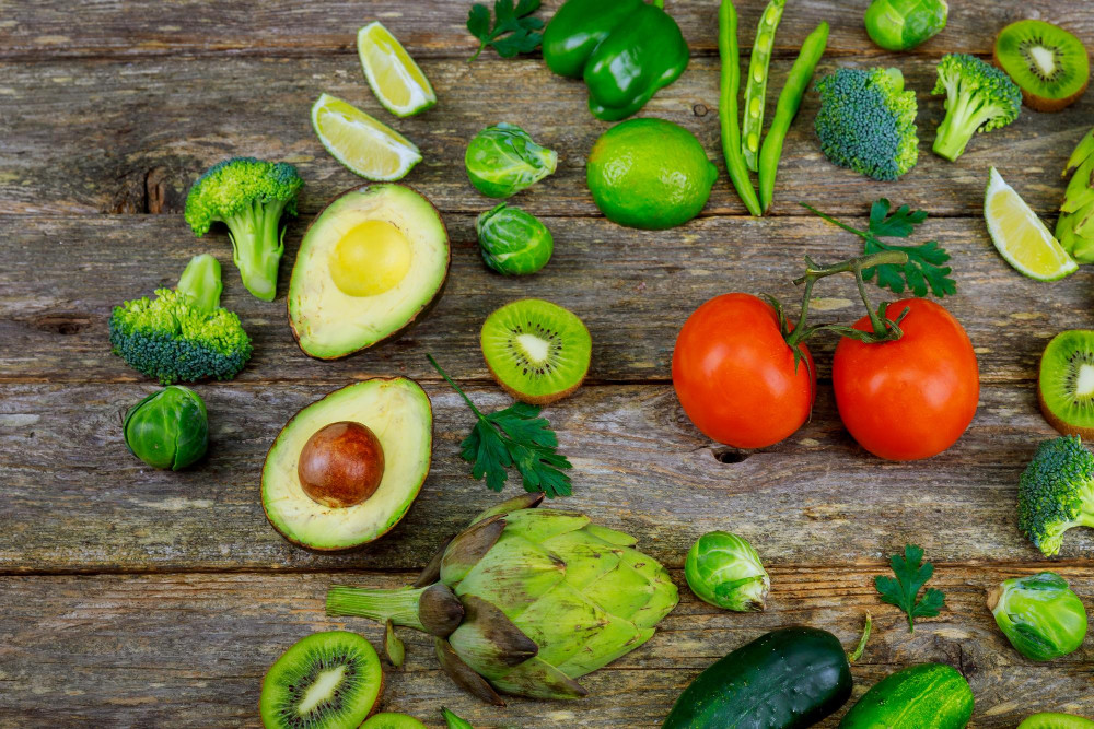 Draufsicht auf einen rustikalen Holztisch mit halbierten Avocados, die zwischen grünem Gemüse wie Brokkoli und Gurken sowie Früchten wie Kiwis und Tomaten liegen.