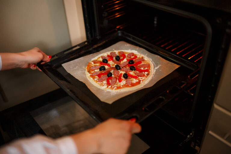 Eine Person schiebt eine belegte Pizza auf einem Backblech in den heißen Backofen.
