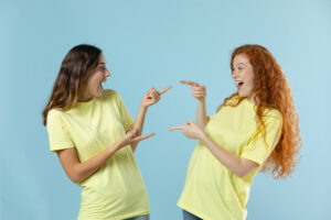 Zwei junge Frauen in gelben T-Shirts zeigen lachend aufeinander vor blauem Hintergrund.