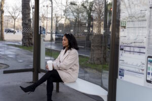 Frau sitzt mit Kaffeebecher an einer verglasten Bushaltestelle und blickt zur Straße.
