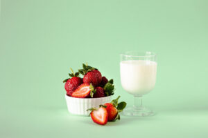 Frische Erdbeeren in einer weißen Schale neben einem Glas Buttermilch auf grünem Hintergrund.