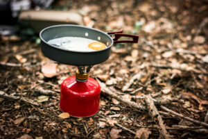 Ein kompakter Campingkocher mit roter Gaskartusche steht auf Waldboden, darauf eine Pfanne mit einem bratenden Spiegelei. Die Szene symbolisiert einfaches, minimalistisches Outdoor-Kochen in der Natur.