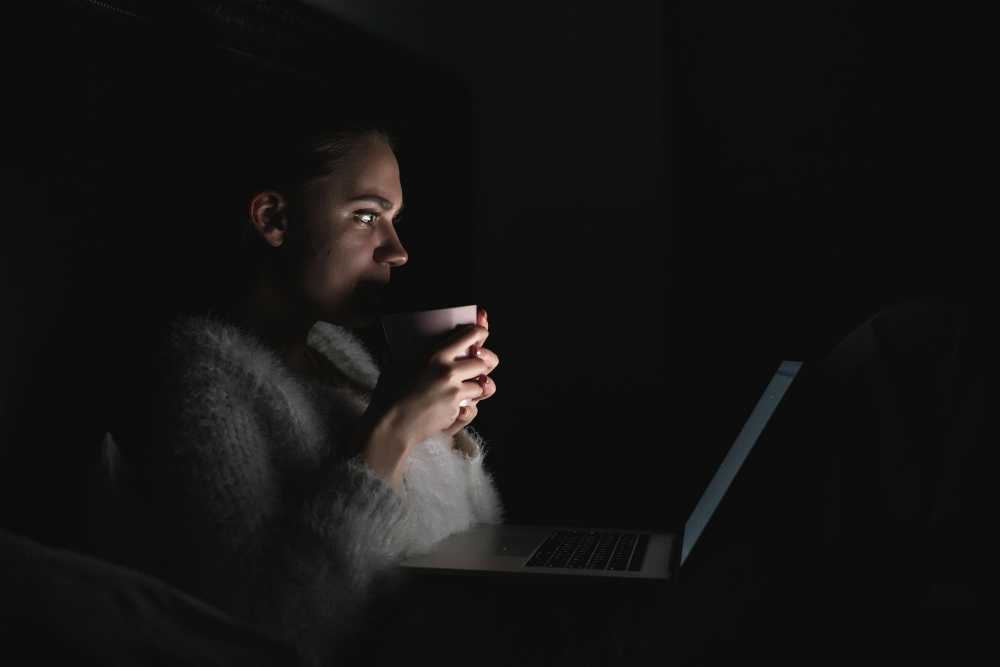 Frau betrachtet im Dunkeln den Laptopbildschirm im Darkmode und hält eine Tasse in der Hand.