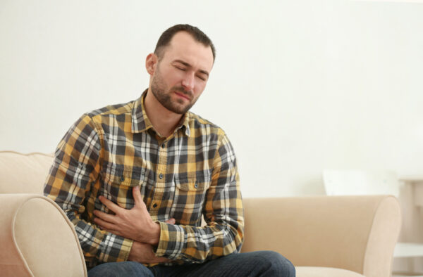 Ein Mann der auf einem Sofa sitzt und sich den Bauch hält als Symbolbild für Ratgeber: Knoblauch im Magen neutralisieren