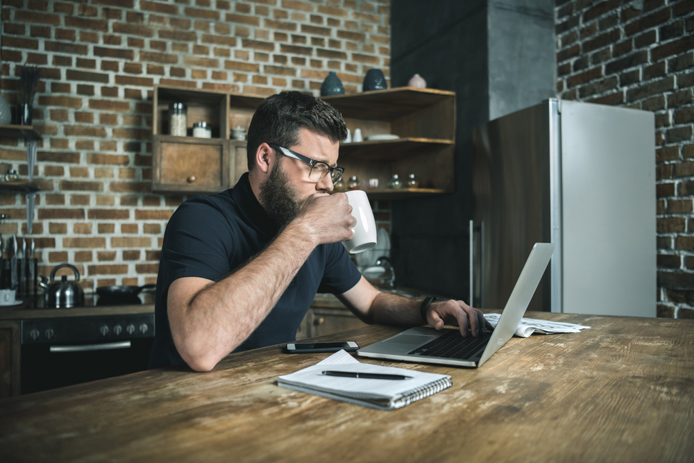 Mann mit Kaffee sitzt am Laptop an einem Holztisch in einem modernen Haushalt als Symbolbild für den Ratgeber: Minijob im Homeoffice