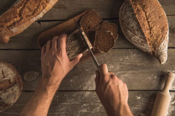 Gesundes dunkles Brot wird von Hand geschnitten als Symbolbild für den Ratgeber: Welches Brot eignet sich am besten zum abnehmen?