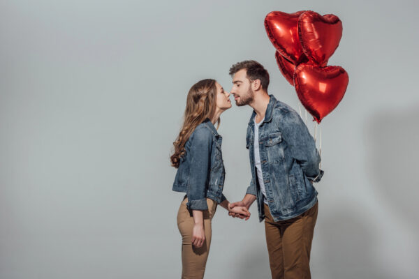 Ein Päärchen hält sich die Hand und küsst sich, während der Mann Herzluftballons hinterm Rücken hält als Symbolbild für den Ratgeber: Last Minute Valentinstagsgeschenk für Verliebte