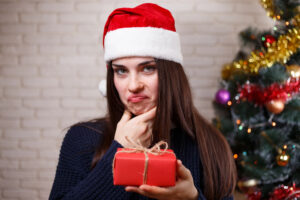 Junge Frau mit Weihnachtsmütze vor einem Weihnachtsbaum hält ein Geschenk in der Hand und schmollt als Symbolbild für den Ratgeber: Wenn Geschenke nicht gefallen