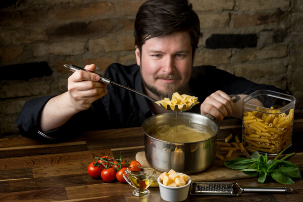 Junger Mann hebt erwartungsvoll eine Nudel mit dem Nudellöffel aus dem Topf als Symbolbild für den Ratgeber: Nudeln kochen leicht gemacht