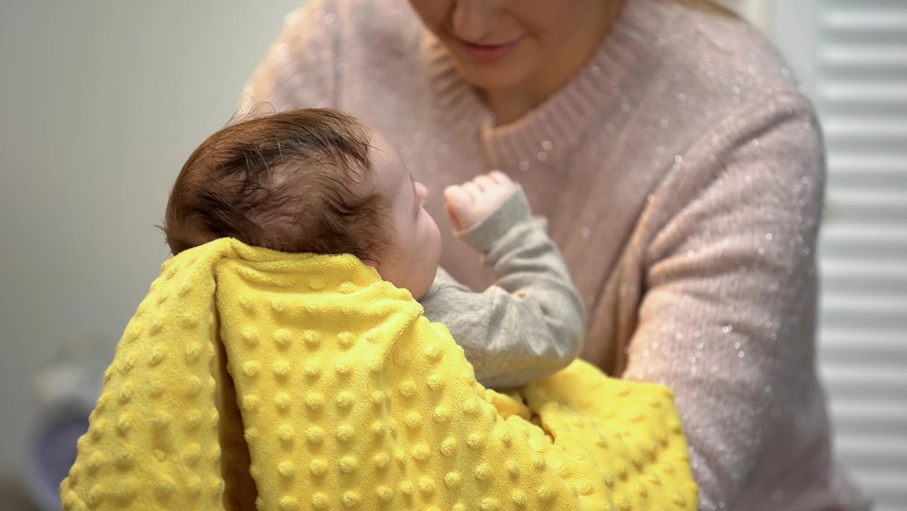 Frischgebackene Mama hält ein Neugeborenes mit einem gelben Spucktuch - Symbolbild für Baby spuckt plötzlich nach jeder Mahlzeit