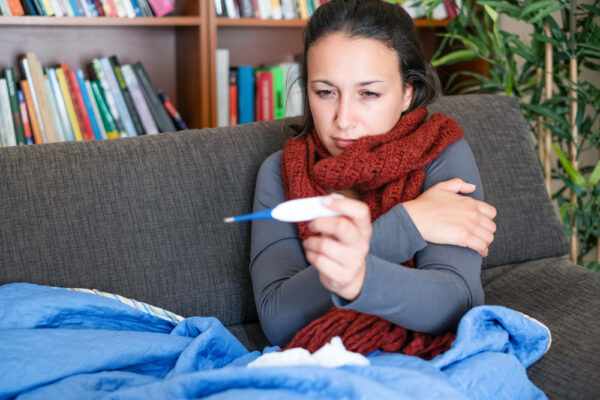 Junge Frau sitzt zusammengekauert mit Schal und Wollpullover auf dem Sofa in eine Decke geschlagen mit einem Fieberthermometer in der Hand als Symbolbild für den Ratgeber: Frieren bei Fieber