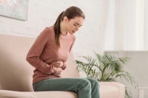 Junge Frau mit Pferdeschwanz sitzt auf dem Sofa, verzieht das Gesicht und hält sich den Bauch als Symbolbild für den Ratgeber: Durchfall nach fettigem Essen