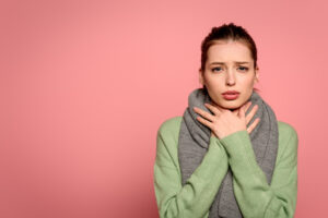Junge Frau mit grauem Schal hält sich den Hals und blickt leidend in die Kamera als Symbolbild für den Ratgeber: Schal bei Halsschmerzen - ja oder nein?