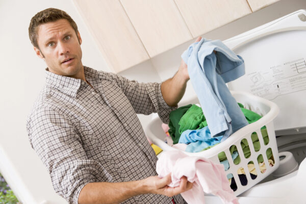 Mann will gerade Wäsche aus dem Wäschekorb herausholen und sieht fragend in die Kamera als Symbolbild für den Beitrag: Warum soll man zwischen Weihnachten und Neujahr keine Wäsche waschen?