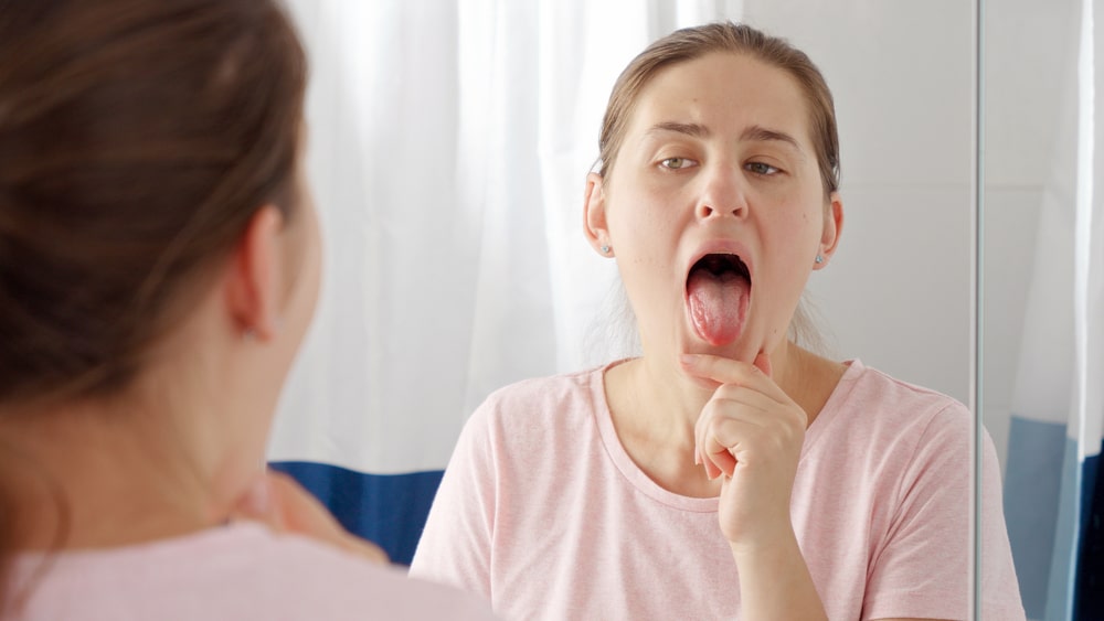 Junge Frau betrachtet ihre Zunge im Spiegel als Symbolbild für den Ratgeber: Bläschen auf der Zunge?