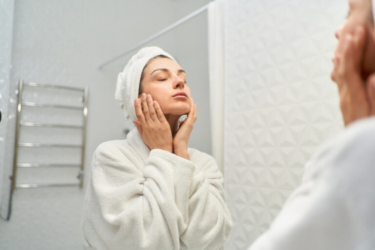 Junge Frau steht mit geschlossenen Augen vor dem Spiegel und berührt ihr Gesicht als Symbolbild für den Ratgeber: Fahle Haut